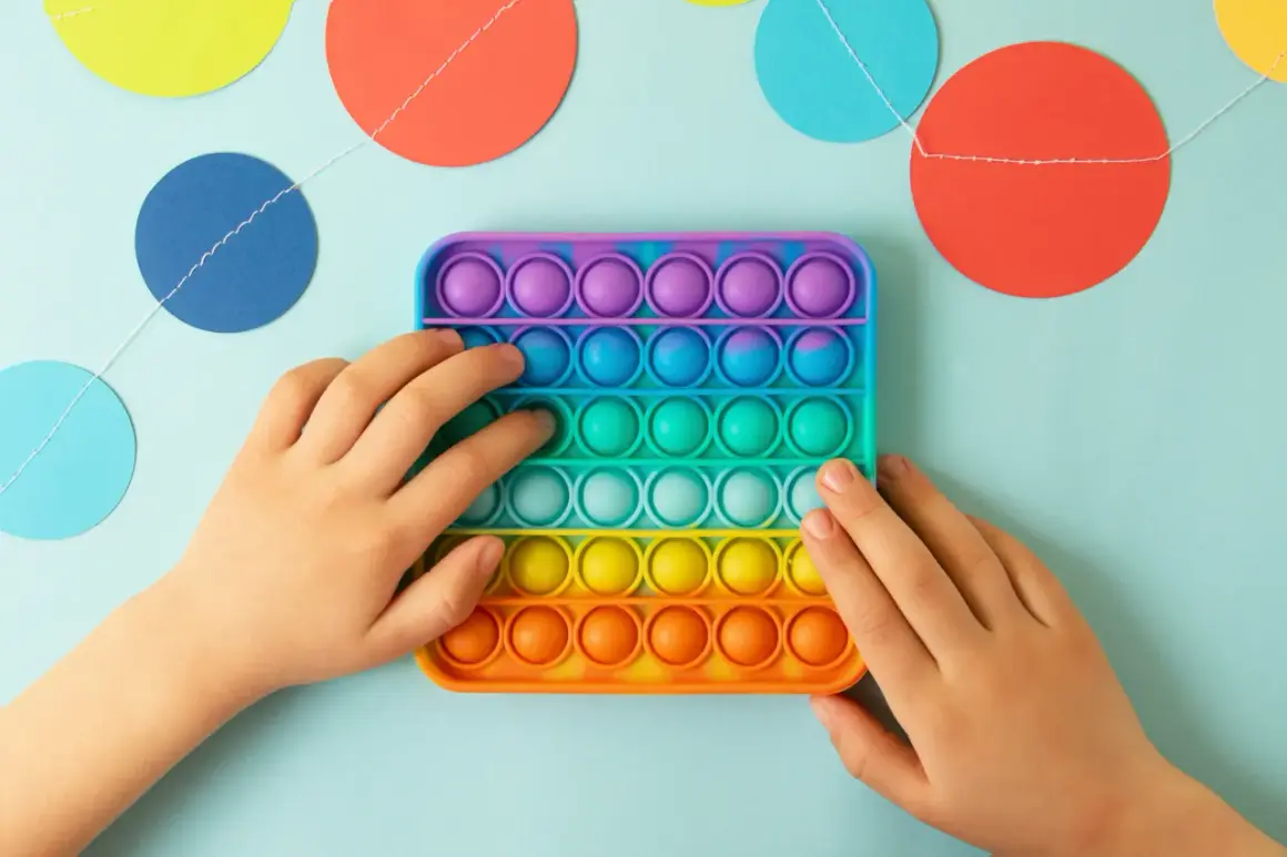 Dziecko bawi się kolorową zabawką sensoryczną typu pop it na jasnoniebieskim tle z girlandą kółek.